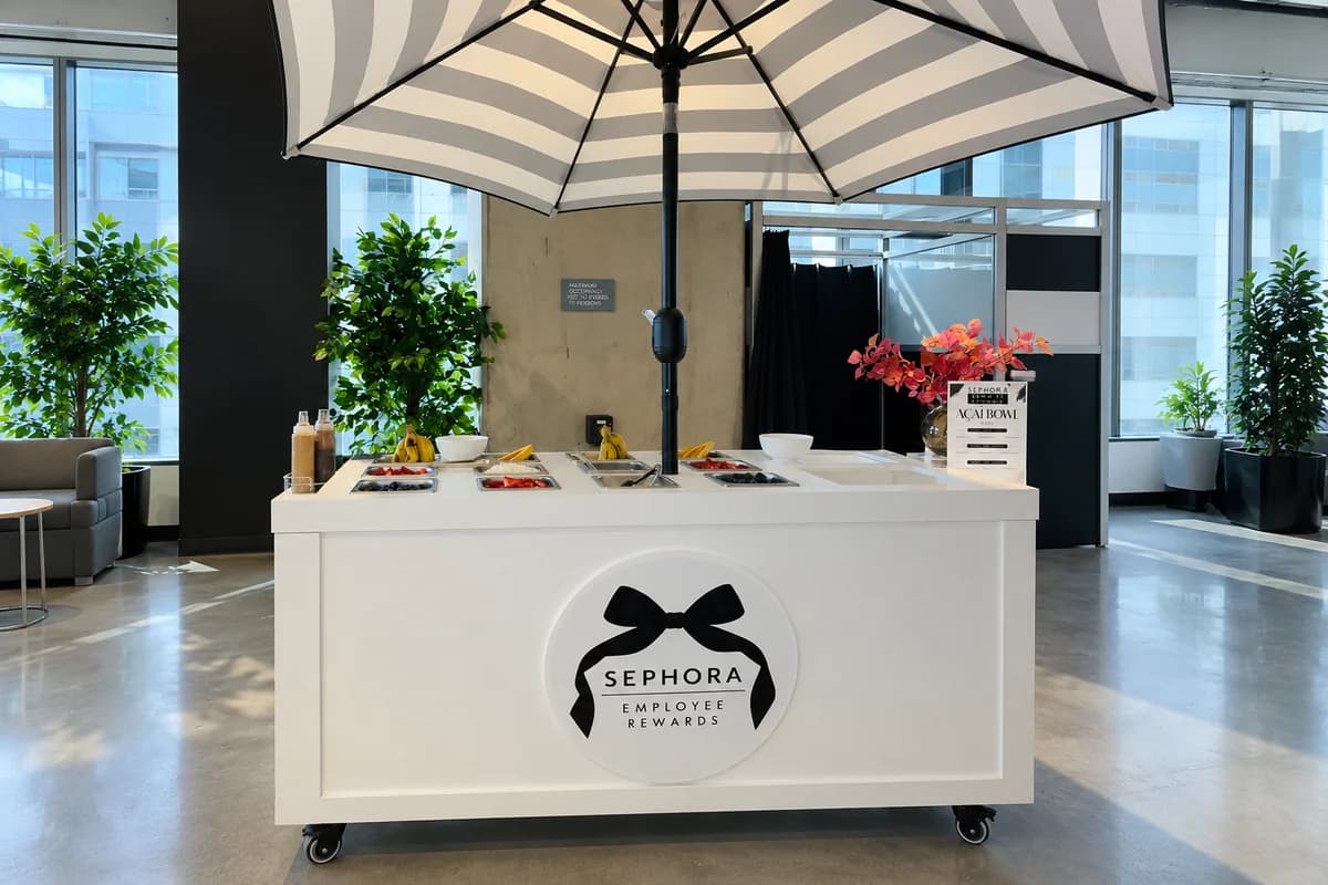 Branded açaí bowl cart with fresh fruit toppings and a striped umbrella at a Sephora employee rewards event