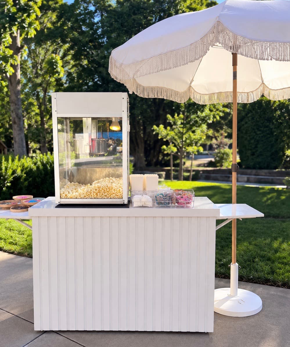 White popcorn cart with a popcorn machine and toppings beneath a fringed umbrella at an outdoor event